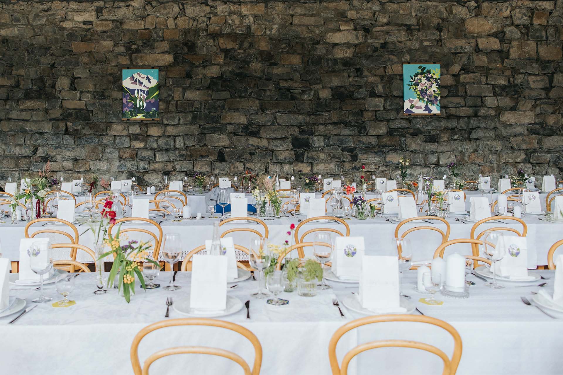 set long tables at a wedding with flowers, coasters and giveaways on them and posters with illustrations on the stone wall behind