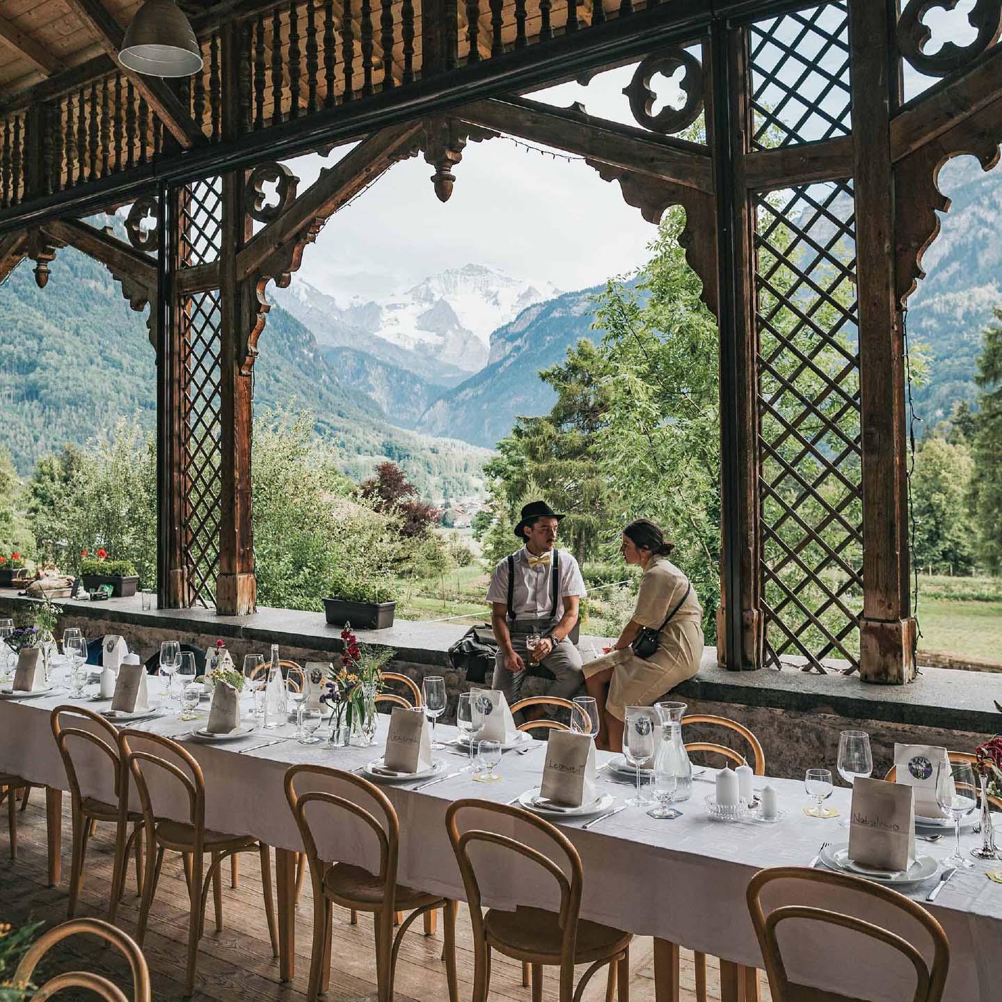 set long tables at a wedding with flowers, coasters and giveaways on them and a couple in the background in front of a swiss mountain scenery